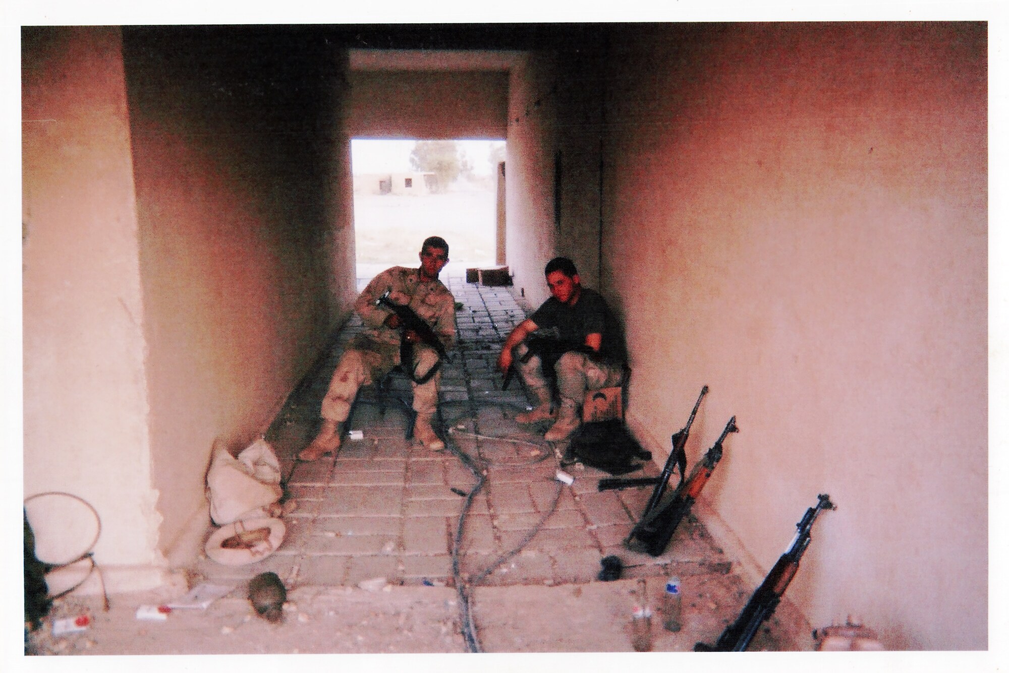 A long tiled corridor inside an Iraqi weapons complex, lined with military packs, bedding, rifles, and personal gear.