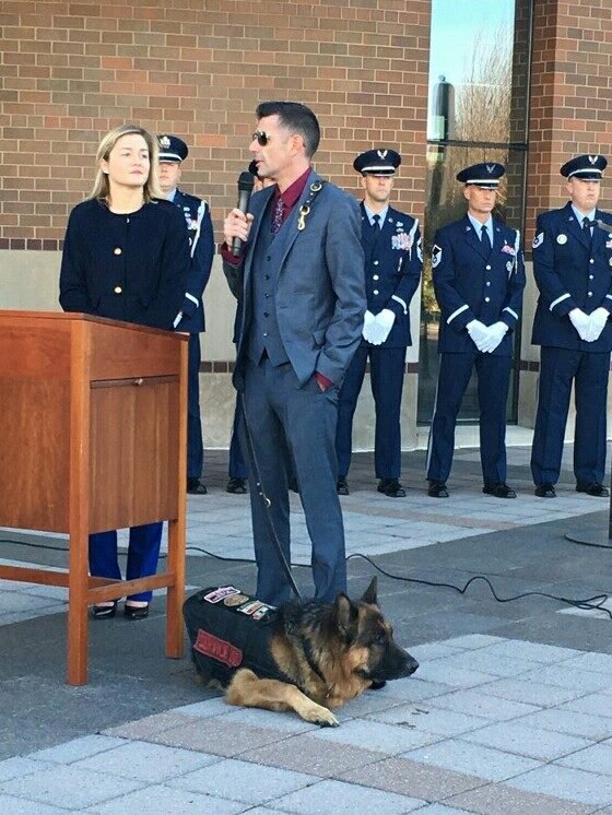 Kenny Bass speaking at a Veterans Day event with Atlas lying beside him.