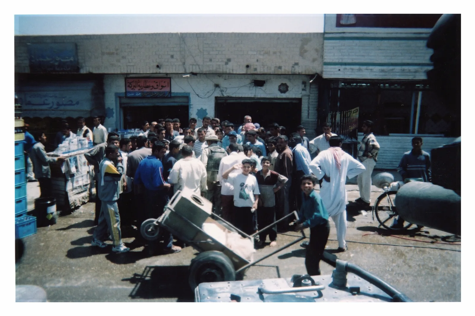 A crowd fills the space before low shops. A helmeted figure stands near the center with his back to the camera. A handcart crosses the foreground diagonally; several faces turn toward the lens.