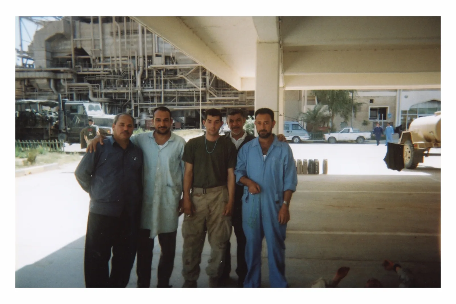 Five men stand together under a broad concrete canopy, some with arms around shoulders. The central man wears a blue beaded loop. Industrial pipes fill the left background; vehicles and open floor extend to the right.