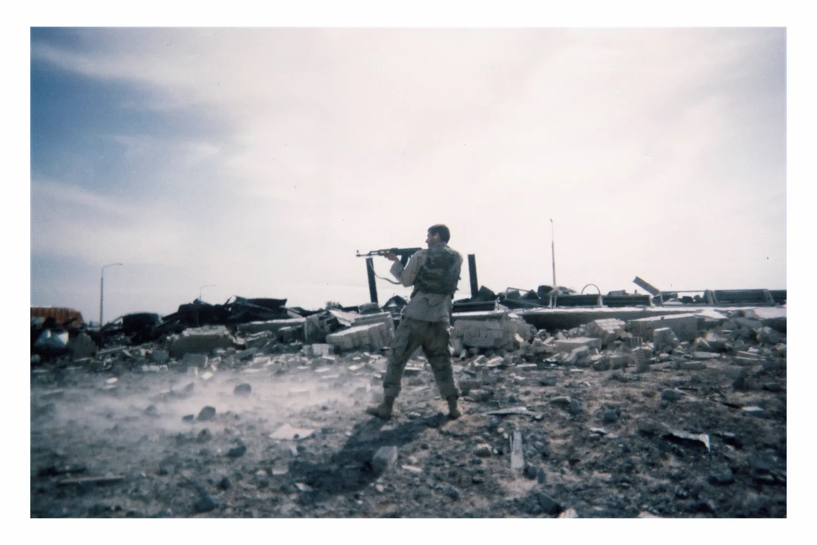 A uniformed man stands wide-legged on broken masonry, his back partly toward the camera. He holds a rifle at shoulder height pointing toward the viewer’s left. Rubble forms a low band beneath a broad sky.