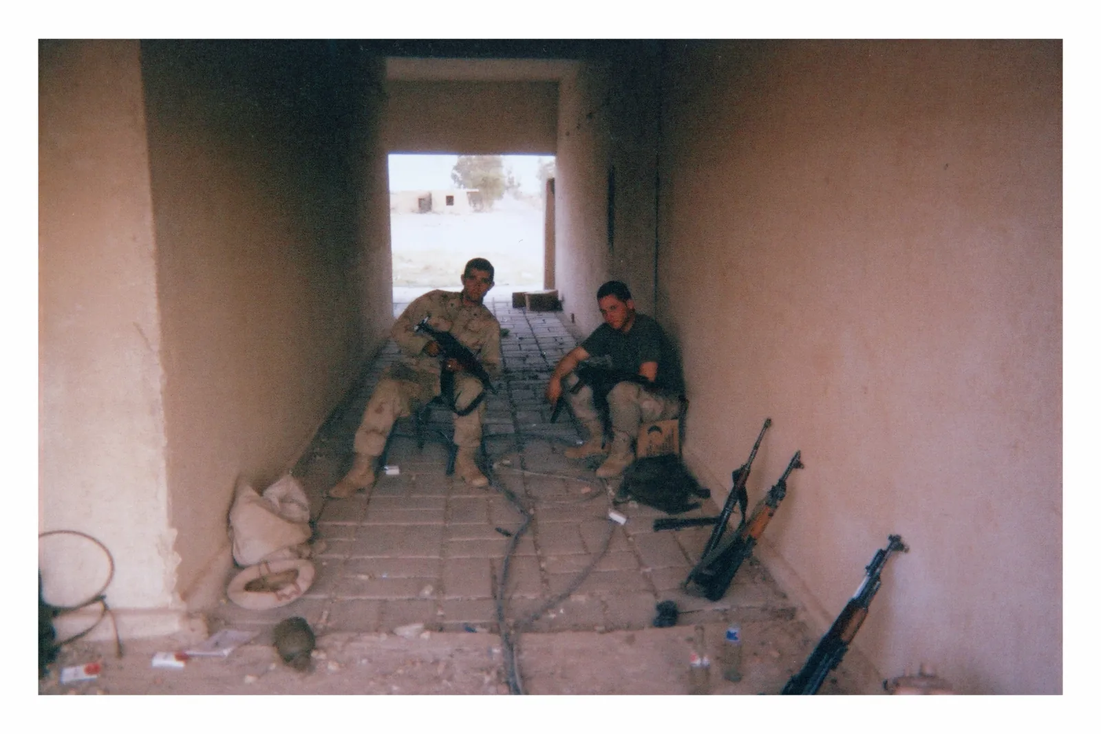 Two men sit near the far end of a narrow tiled hallway. One is near the center; the other is to his right in the photograph, against the wall. Rifles lean along that right wall.
