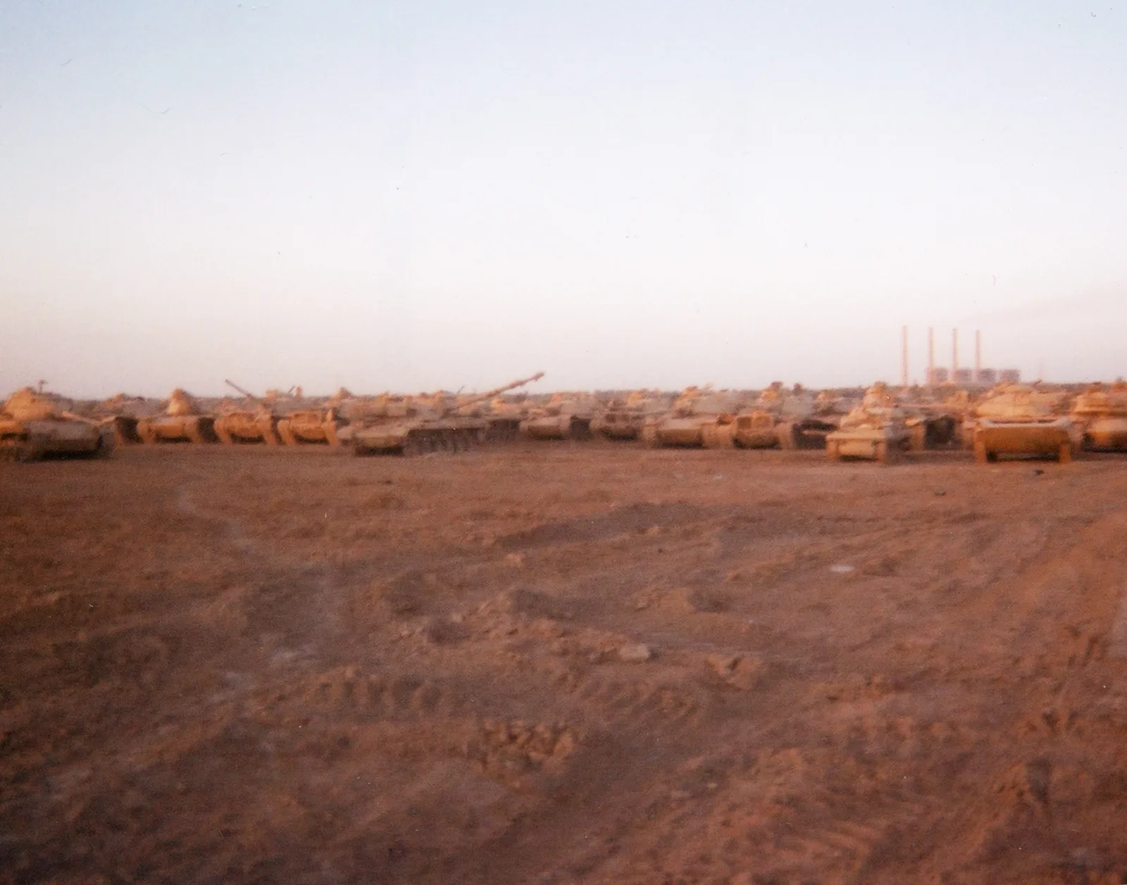 A low line of tanks stretches across tracked earth. Curving ruts fill the broad foreground, and a pale sky fills the upper half. Four thin industrial stacks rise near the right horizon.