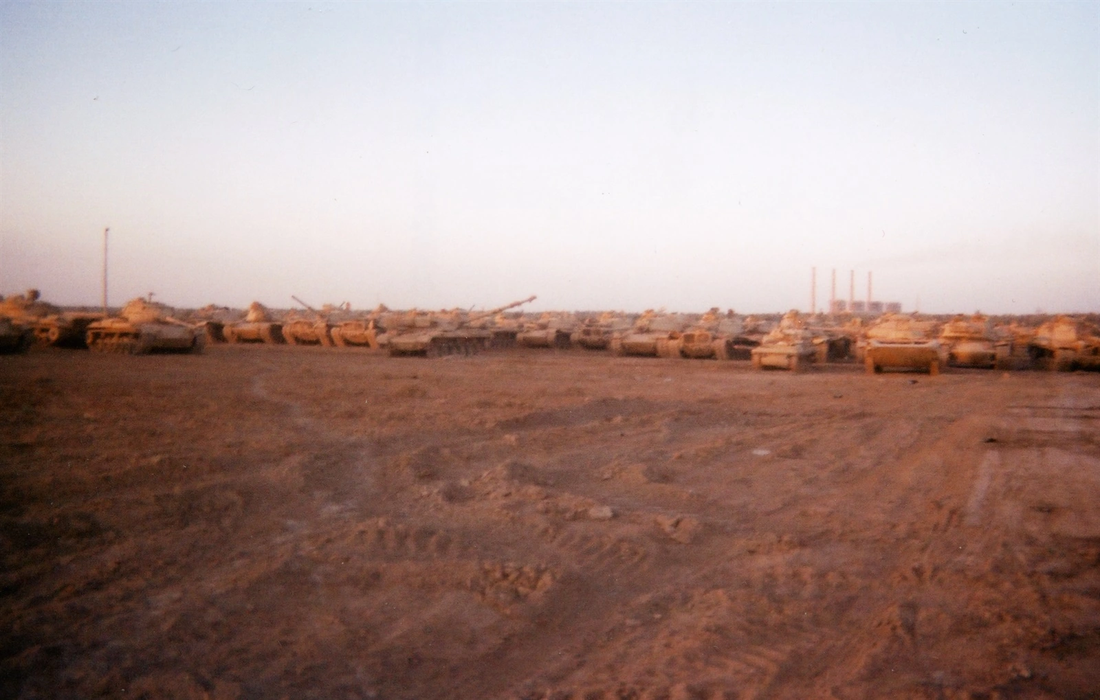 A low line of tanks stretches across tracked earth. Curving ruts fill the broad foreground, and a pale sky fills the upper half. A thin pole stands toward the left, and four industrial stacks rise near the right horizon.