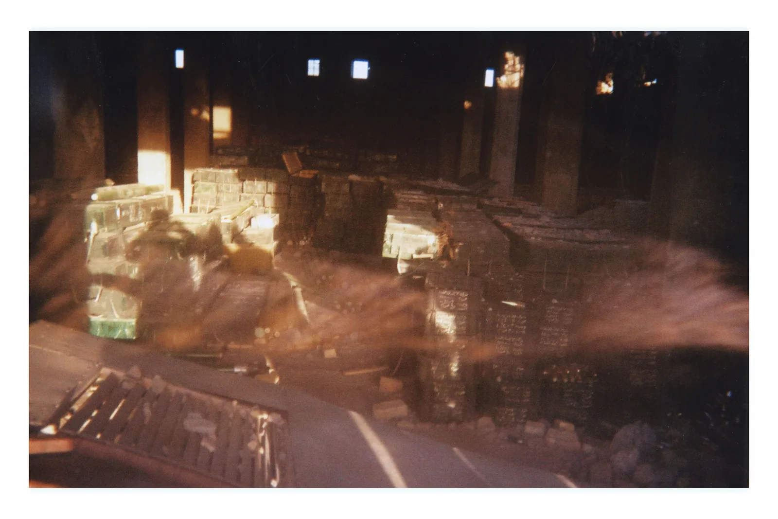 Stacks of boxes occupy a dark interior divided by heavy columns. Small high openings brighten the rear wall. Broad warm streaks pass across the scene, blurring and partly obscuring the containers.