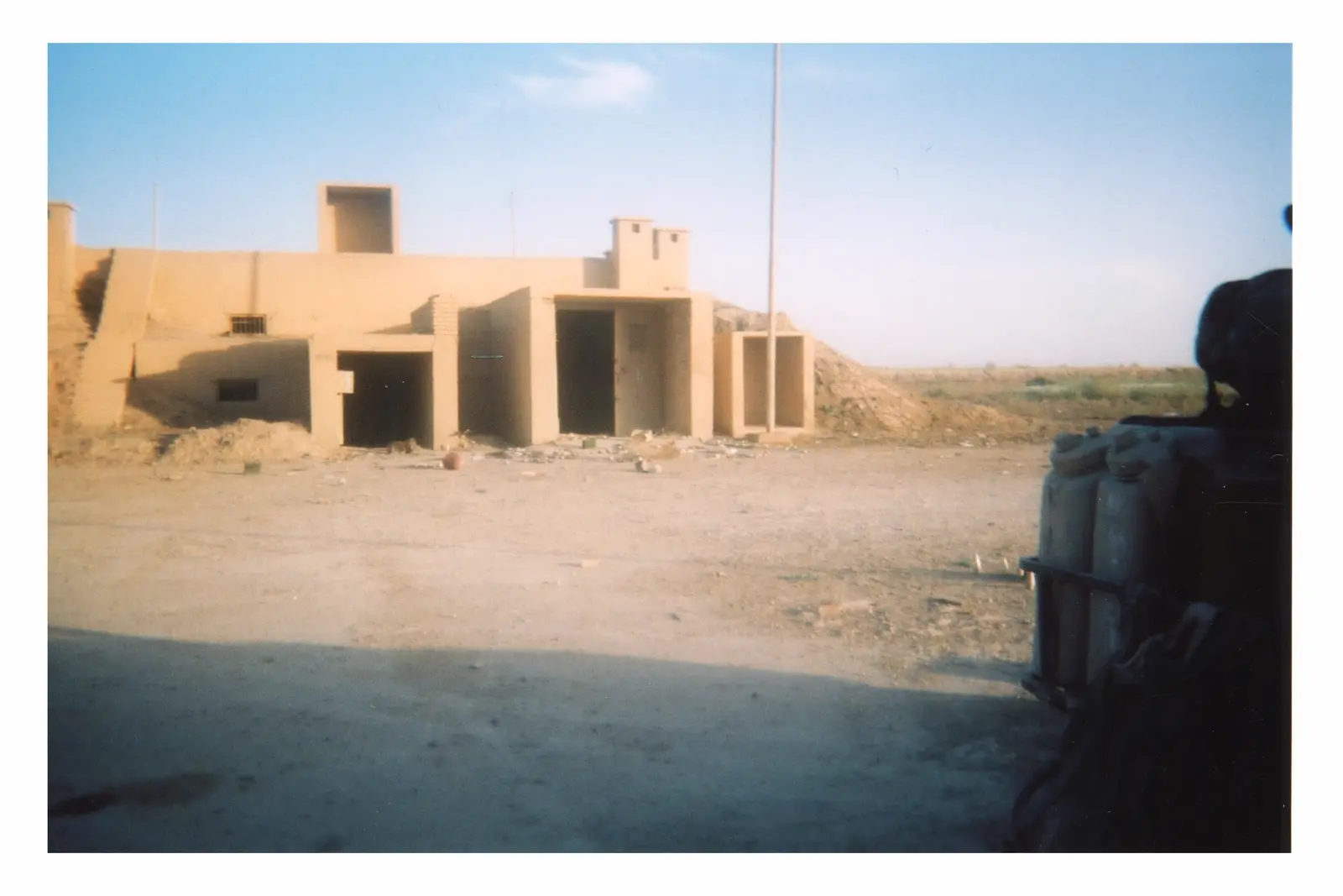A low tan concrete bunker with thick projecting entrance bays and dark openings stands in a bare yard, with earth banked around its rear and right side.