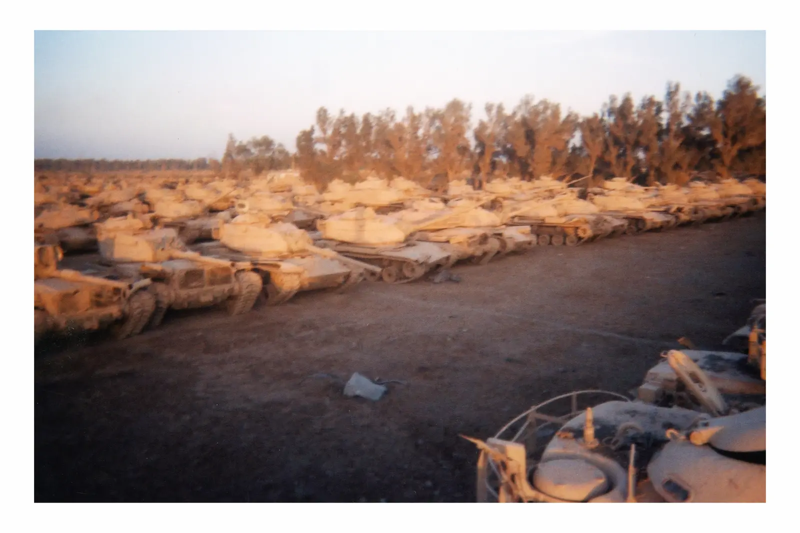 Dense diagonal rows of pale tanks and wheeled armored vehicles fill a dusty field, with trees forming a dark band along the horizon.