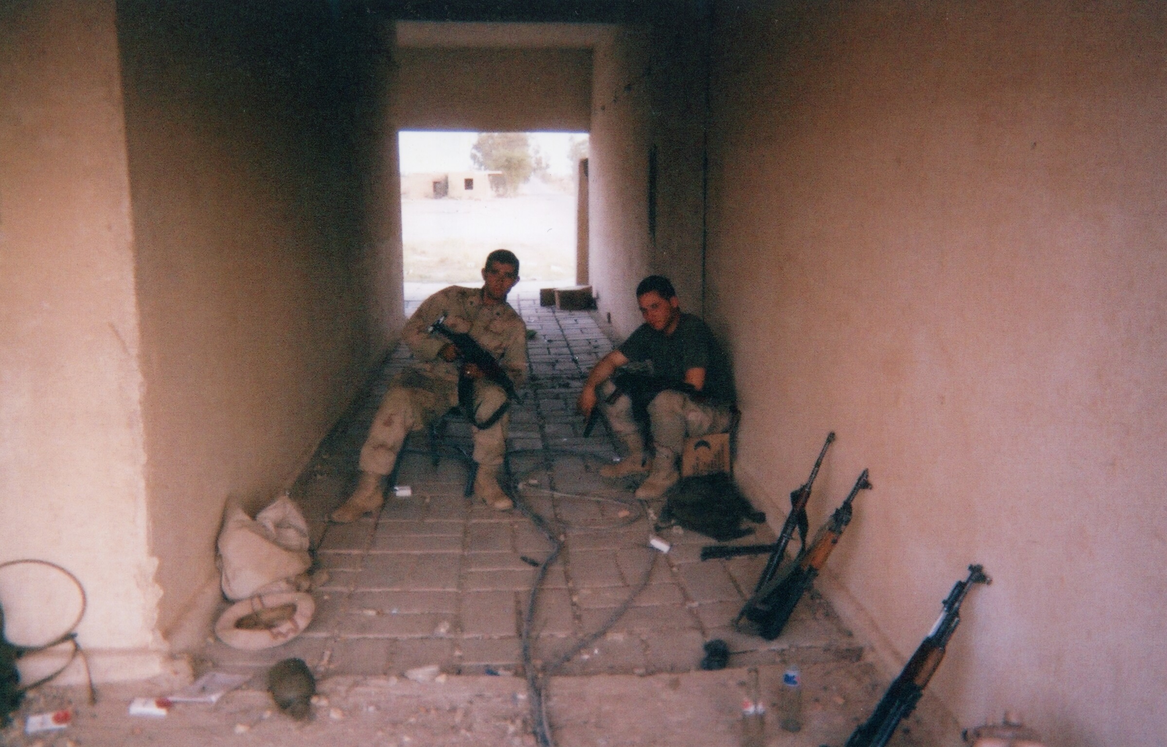 Two Marines in desert utilities resting on the tiled floor of a narrow corridor, rifles and packs beside them, daylight at the far opening.