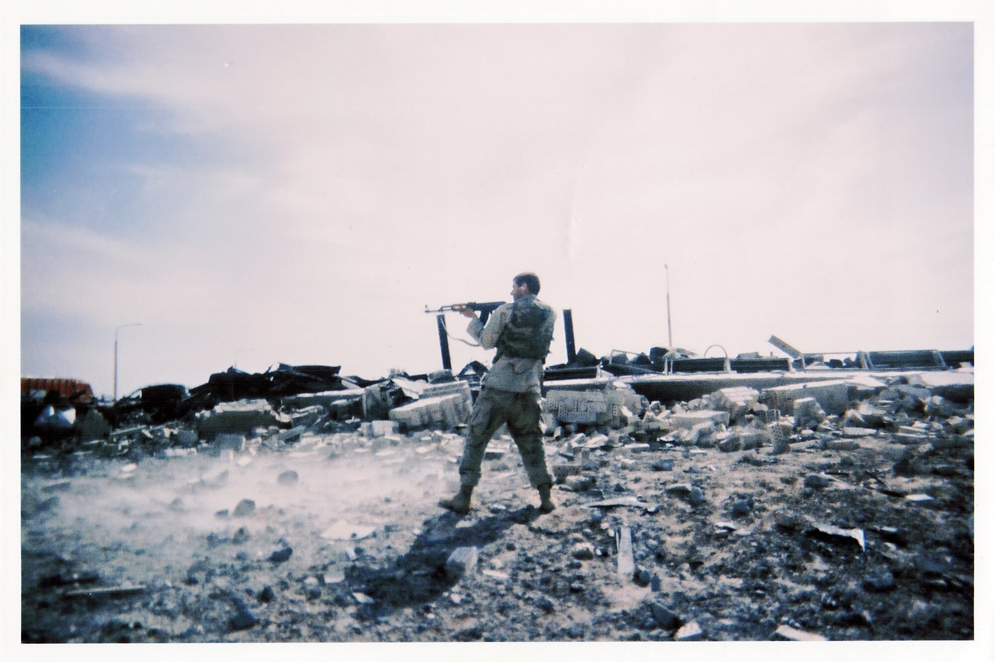 Kenny Bass standing amid rubble and firing an AK toward disabled vehicles inside an Iraqi weapons complex in 2003.