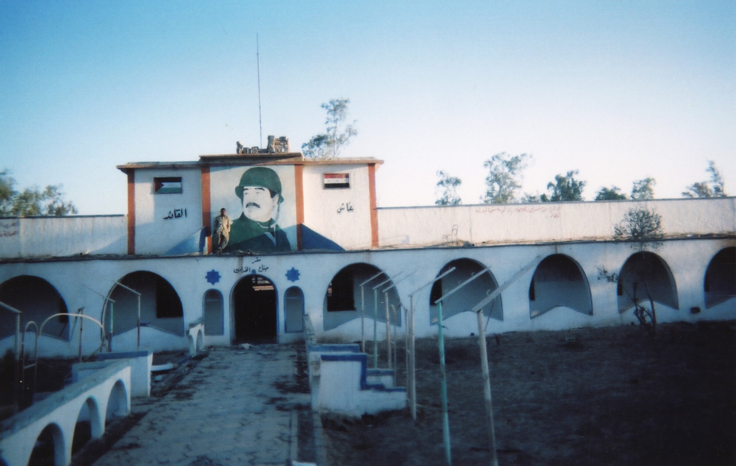 A low arcaded barracks courtyard with a painted mural of a uniformed figure and an Iraqi flag above the central entrance.
