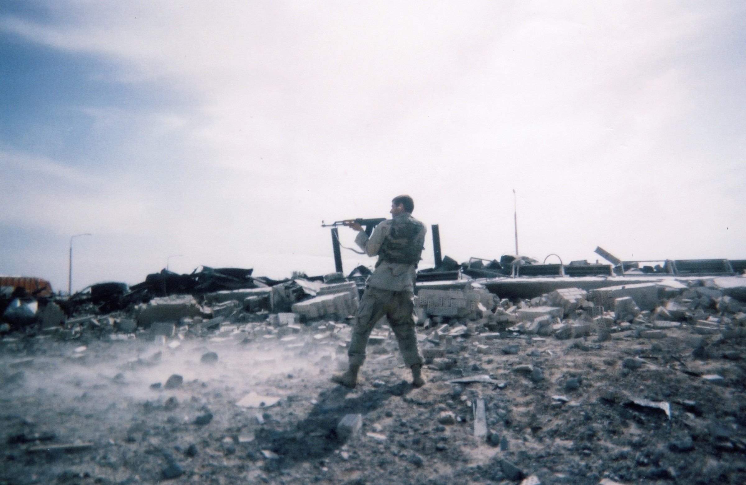 A Marine standing in extensive rubble and firing an AK-style rifle toward damaged vehicles in the distance.