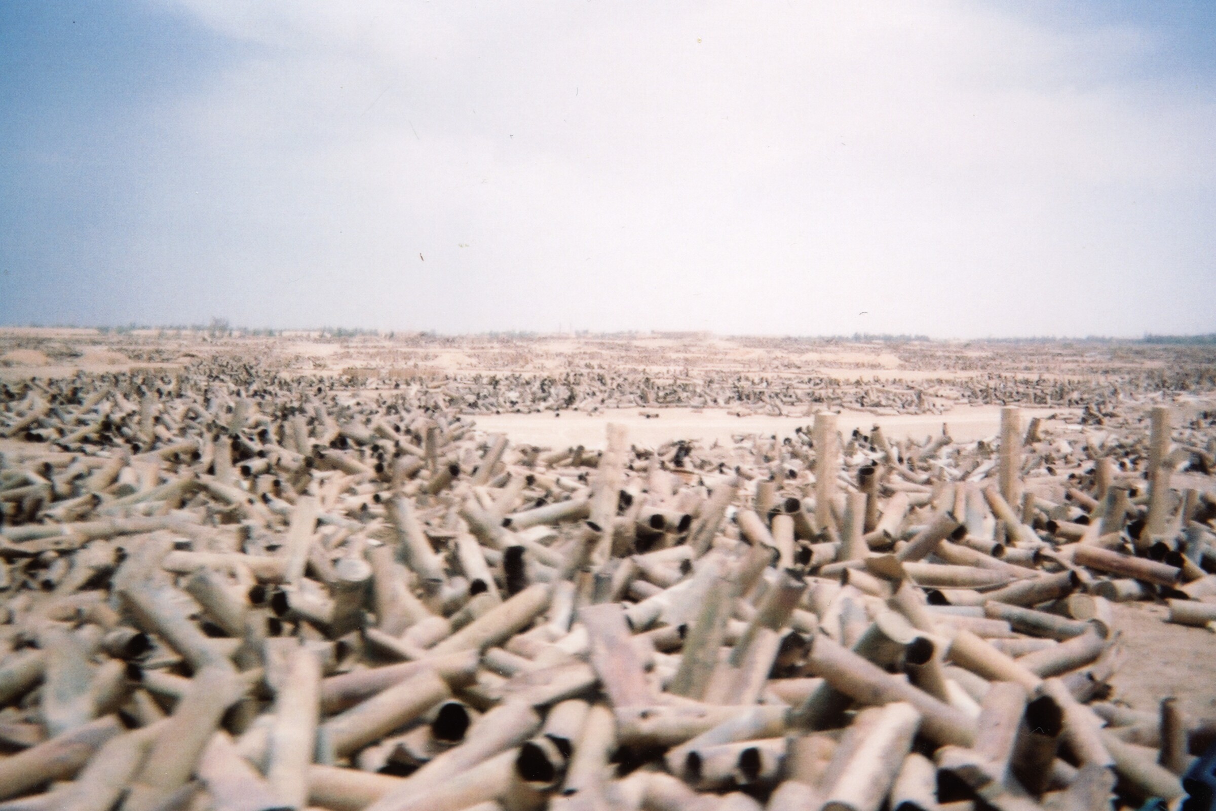 A vast Iraqi desert landscape covered with thousands of spent artillery shell casings beneath a pale blue sky.