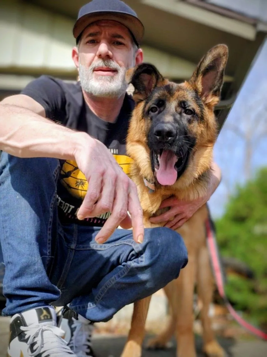 Kenny Bass crouches outdoors beside Arlo, a young German Shepherd.