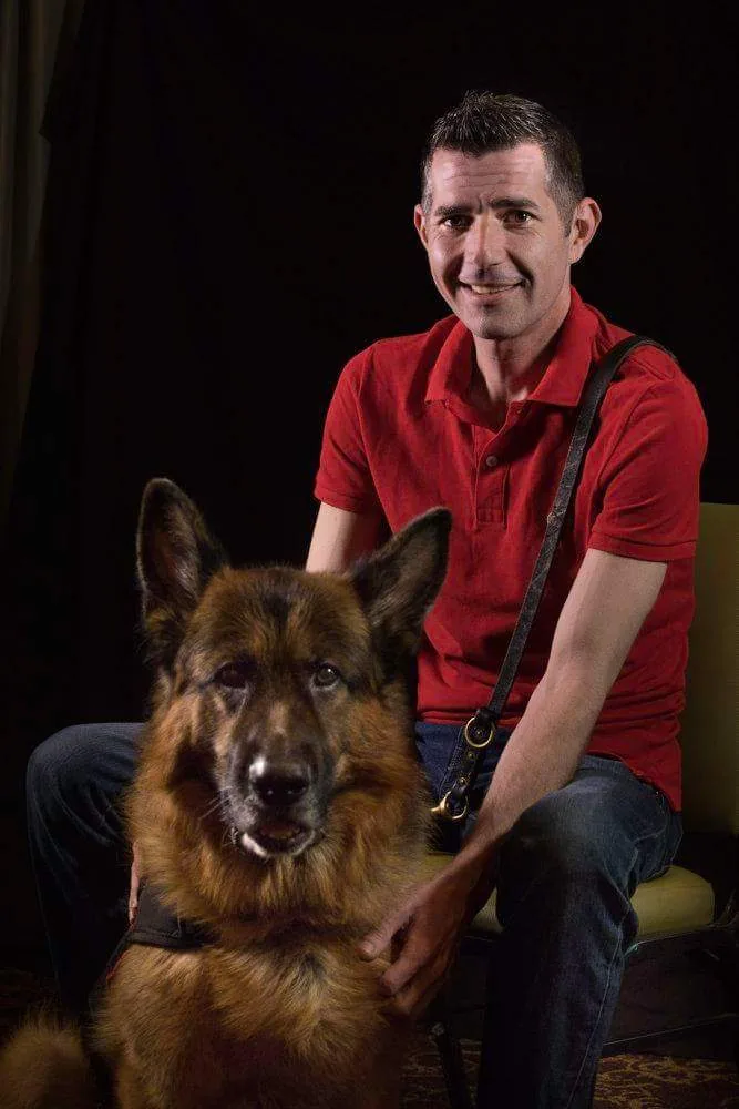 Kenny Bass sits beside Atlas against a dark background, wearing a red polo shirt.