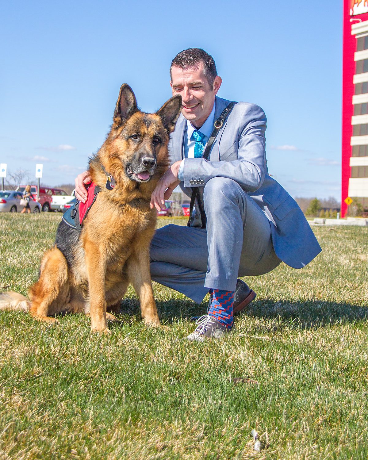 Kenny Bass kneeling beside Atlas, a German Shepherd service dog, outdoors.