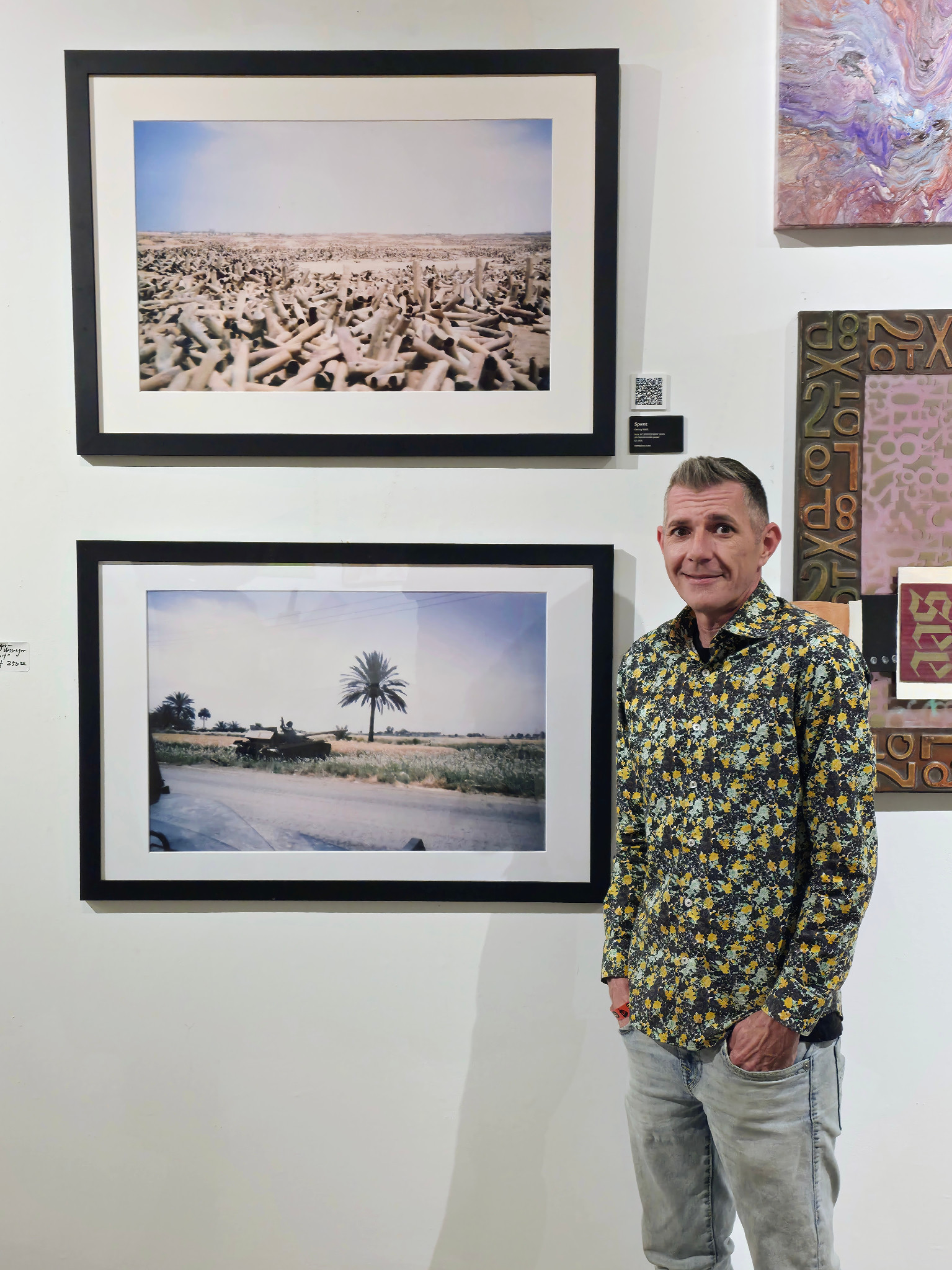 Kenny Bass standing beside framed prints of Spent, above, and West of Eden, below, on a gallery wall.