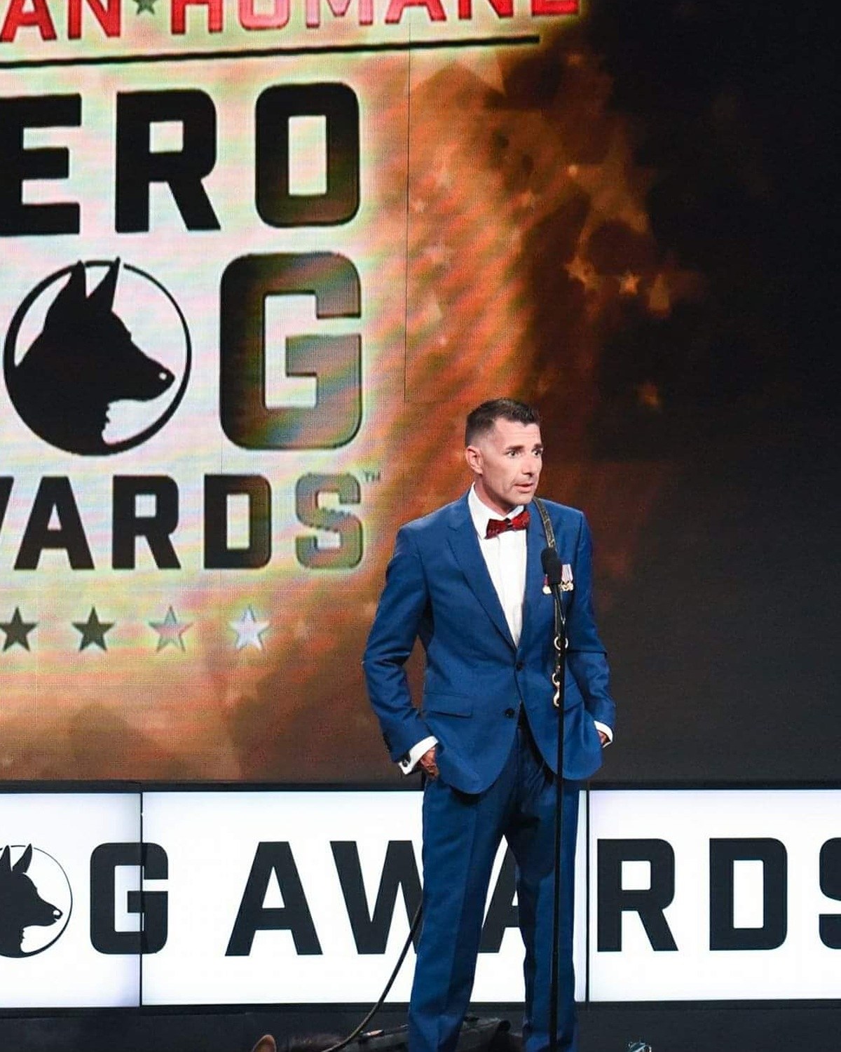 Kenny Bass speaking at a microphone on the American Humane Hero Dog Awards stage with Atlas lying beside him.