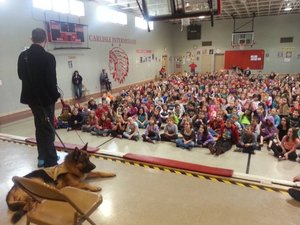 Kenny Bass and Atlas seen from the stage while speaking to a large group of students seated in a school gymnasium.