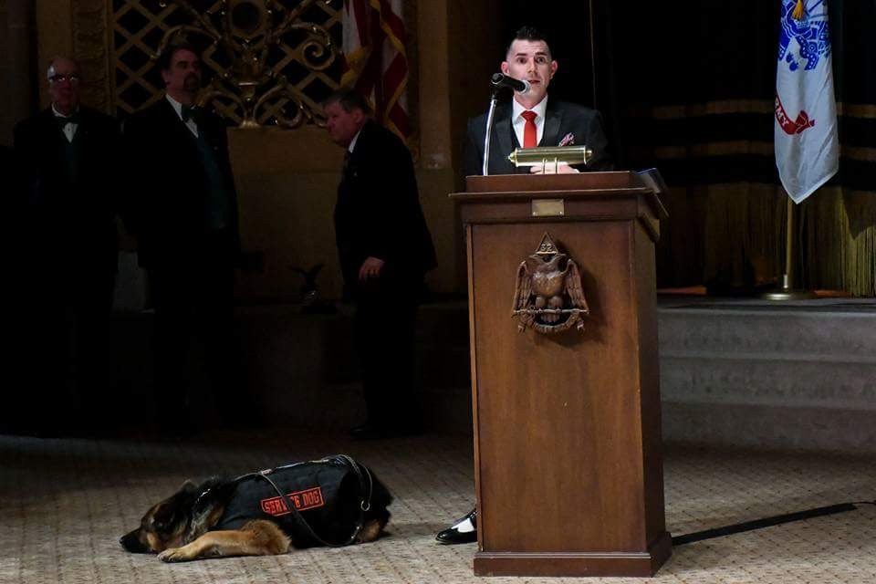 Kenny Bass speaking from a formal podium while Atlas rests beside him in his service-dog vest.