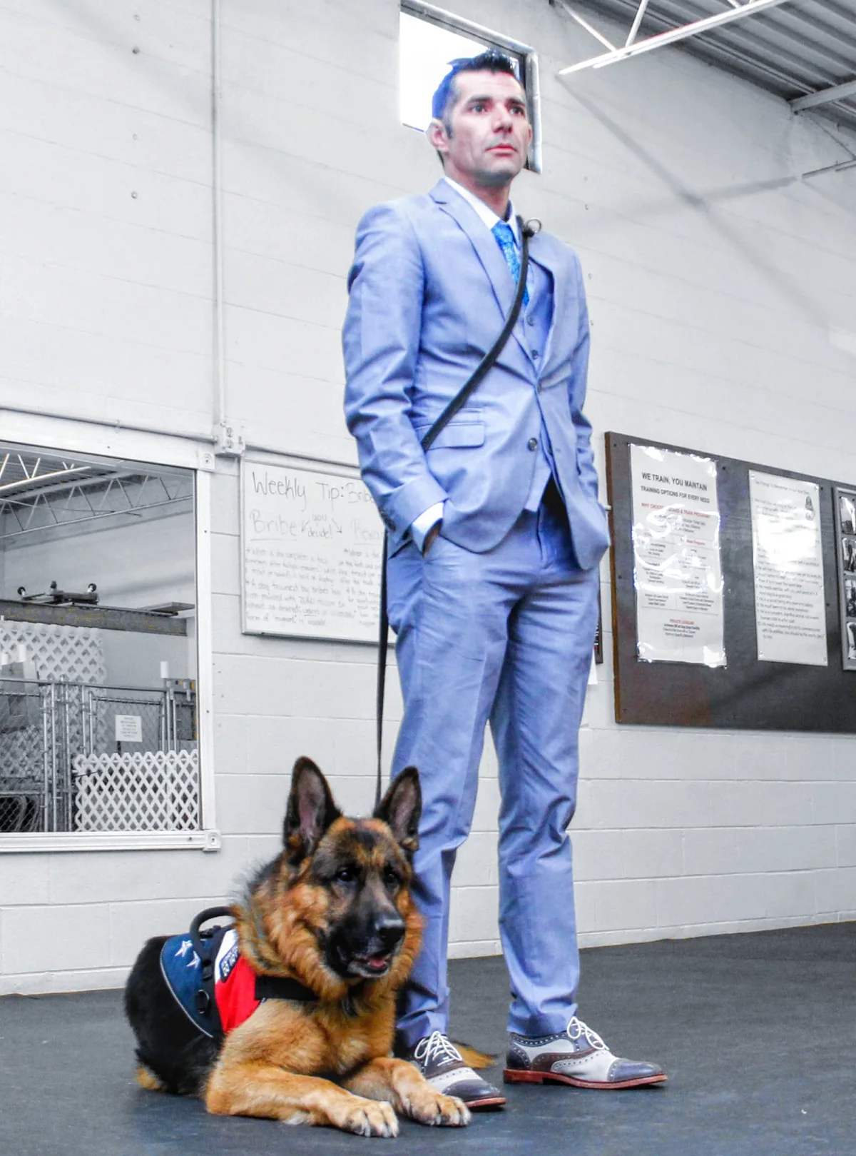 Kenny Bass standing in a light blue suit, with Atlas lying at his feet inside a training facility.