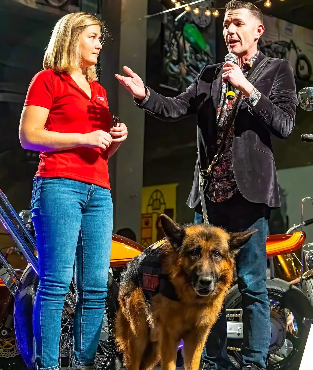 Chrissy Faulkner in a red shirt beside Kenny Bass, speaking into a microphone and gesturing, with Atlas in the foreground.